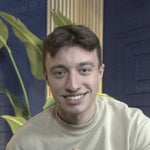 Smiling young man in a light-colored sweatshirt against a blue background with a green plant.