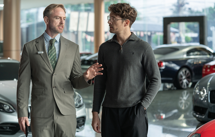 A salesman wears a silver Plaud NotePin on his lapel while speaking with a client in a car dealership.