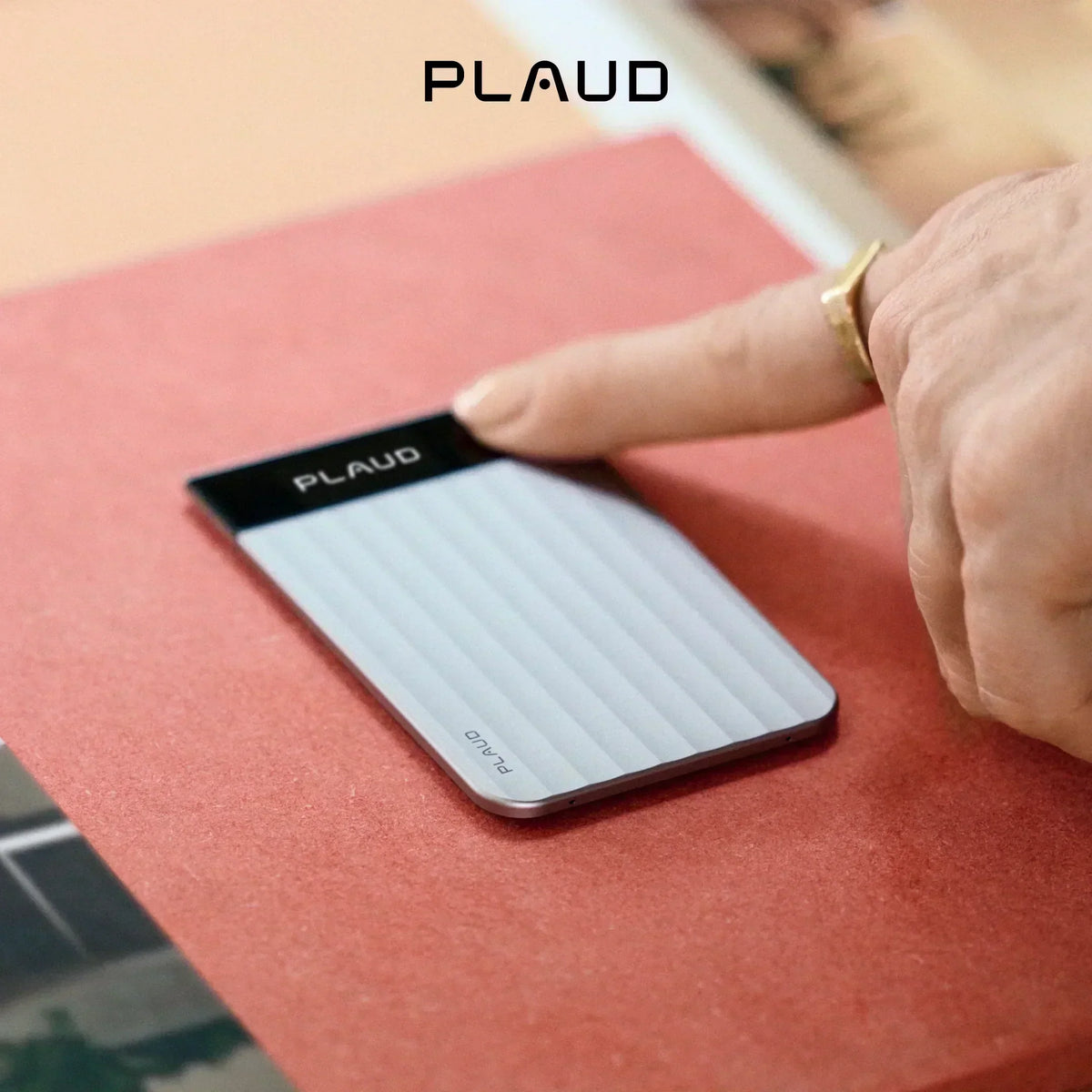 A finger touches the screen of a silver, ribbed Plaud Note Pro AI voice recorder on a red surface.