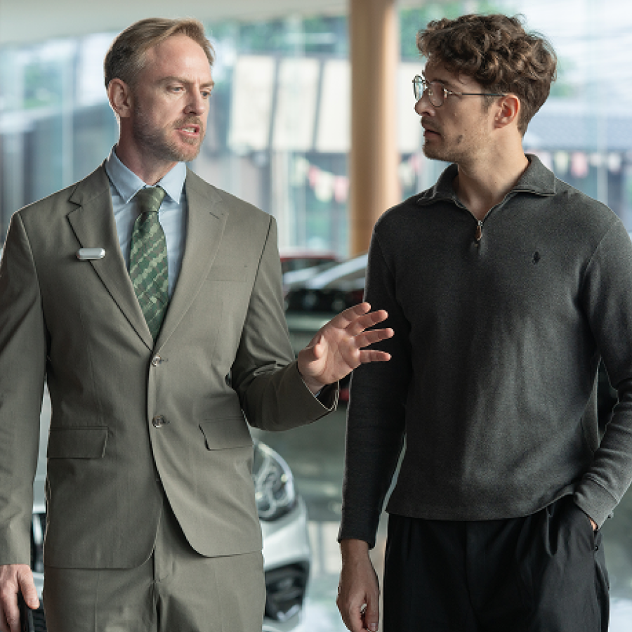 A man in a suit wearing a silver Plaud NotePin on his lapel speaks with another man in a showroom.