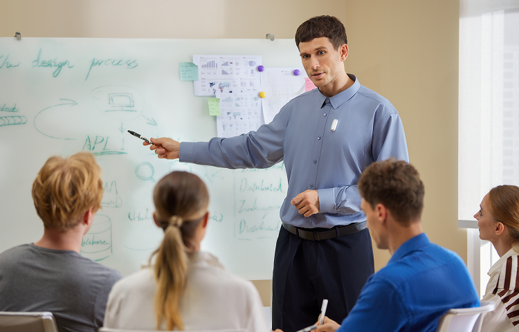 Man in a blue shirt wearing a silver Plaud NotePin presents to colleagues at a whiteboard.