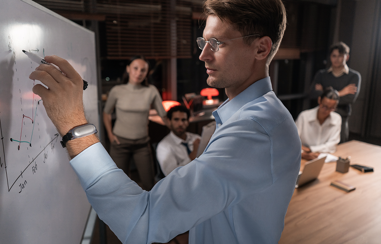 Man wearing a silver Plaud NotePin on a wristband draws a graph on a whiteboard during a team meeting.