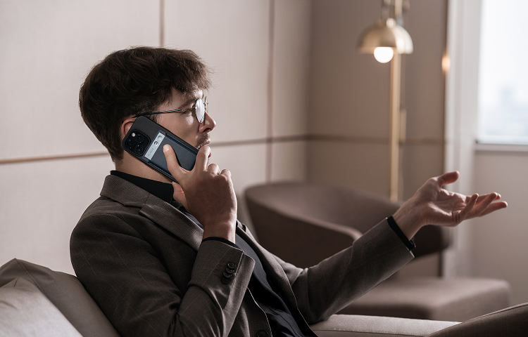 A man in a suit records a phone call using a black Plaud Note attached to the back of his smartphone.