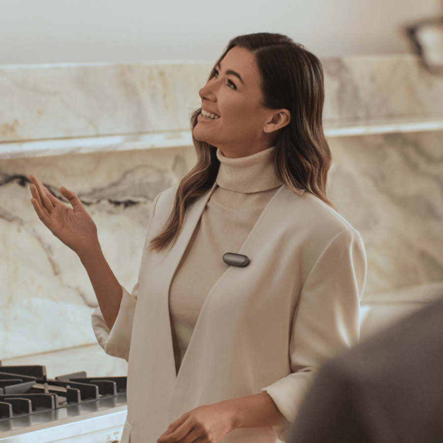 Woman wearing a Cosmic Gray Plaud NotePin on her white blazer lapel while gesturing and smiling.