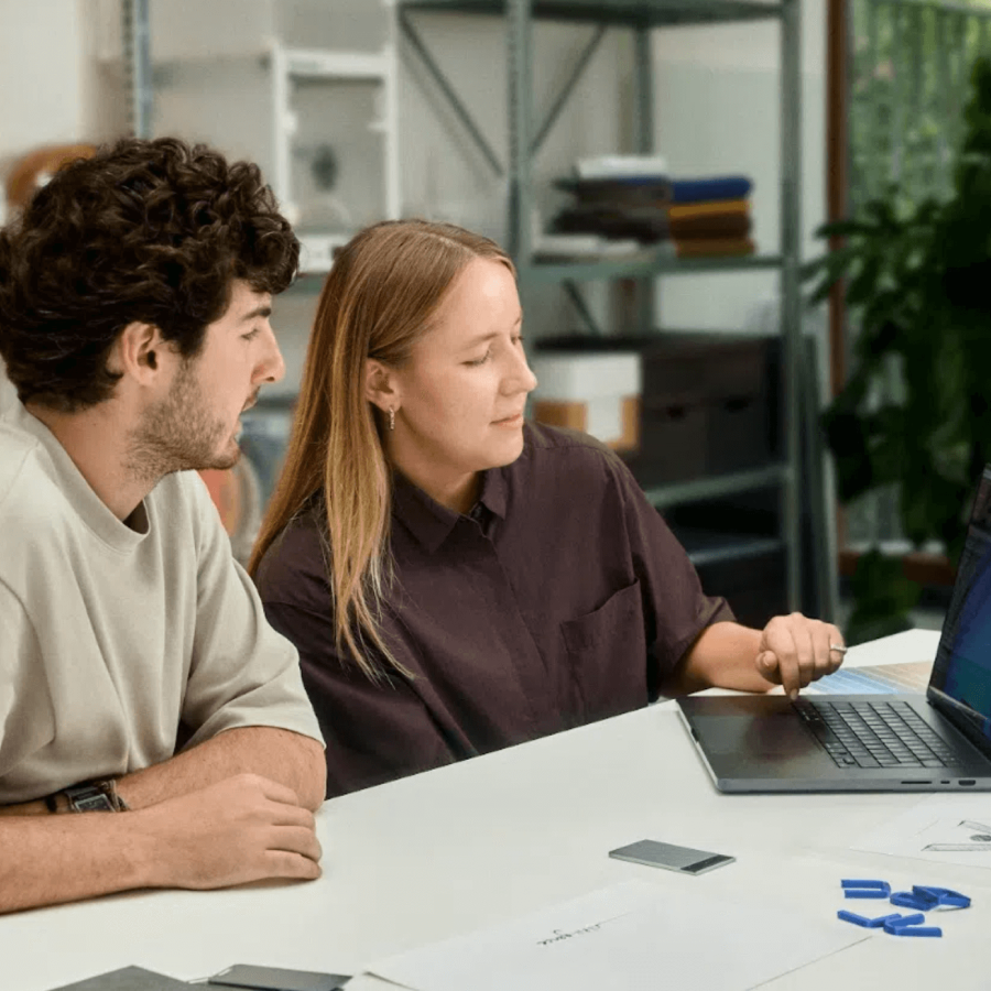 Two colleagues collaborating at a desk with a silver Plaud Note AI voice recorder nearby.