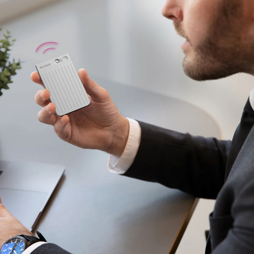 Man in a suit speaking into a silver Plaud Note Pro AI voice recorder while sitting at an office desk.