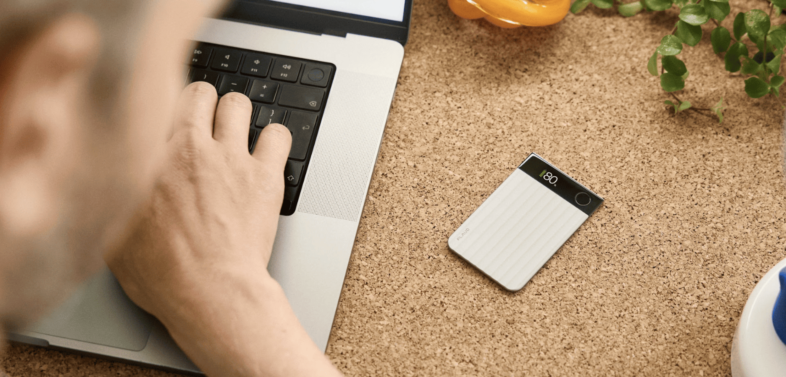 Silver Plaud Note Pro on a cork desk next to a person typing on a laptop.