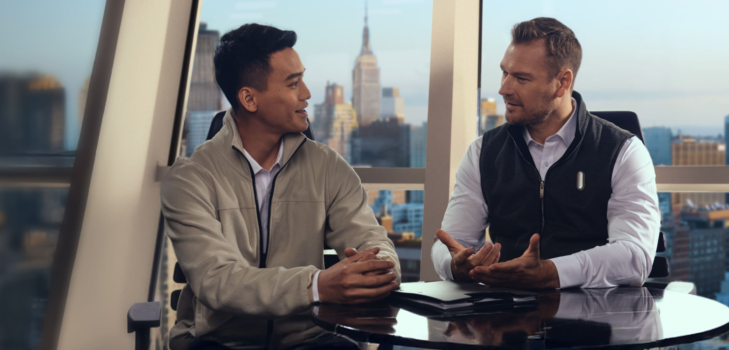 Two men in a meeting with a city view. One wears a silver Plaud NotePin on his vest.