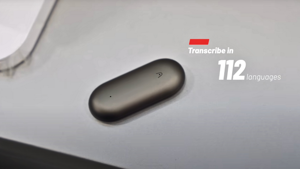 Dark gray Plaud NotePin on a desk with text stating it transcribes in 112 languages.