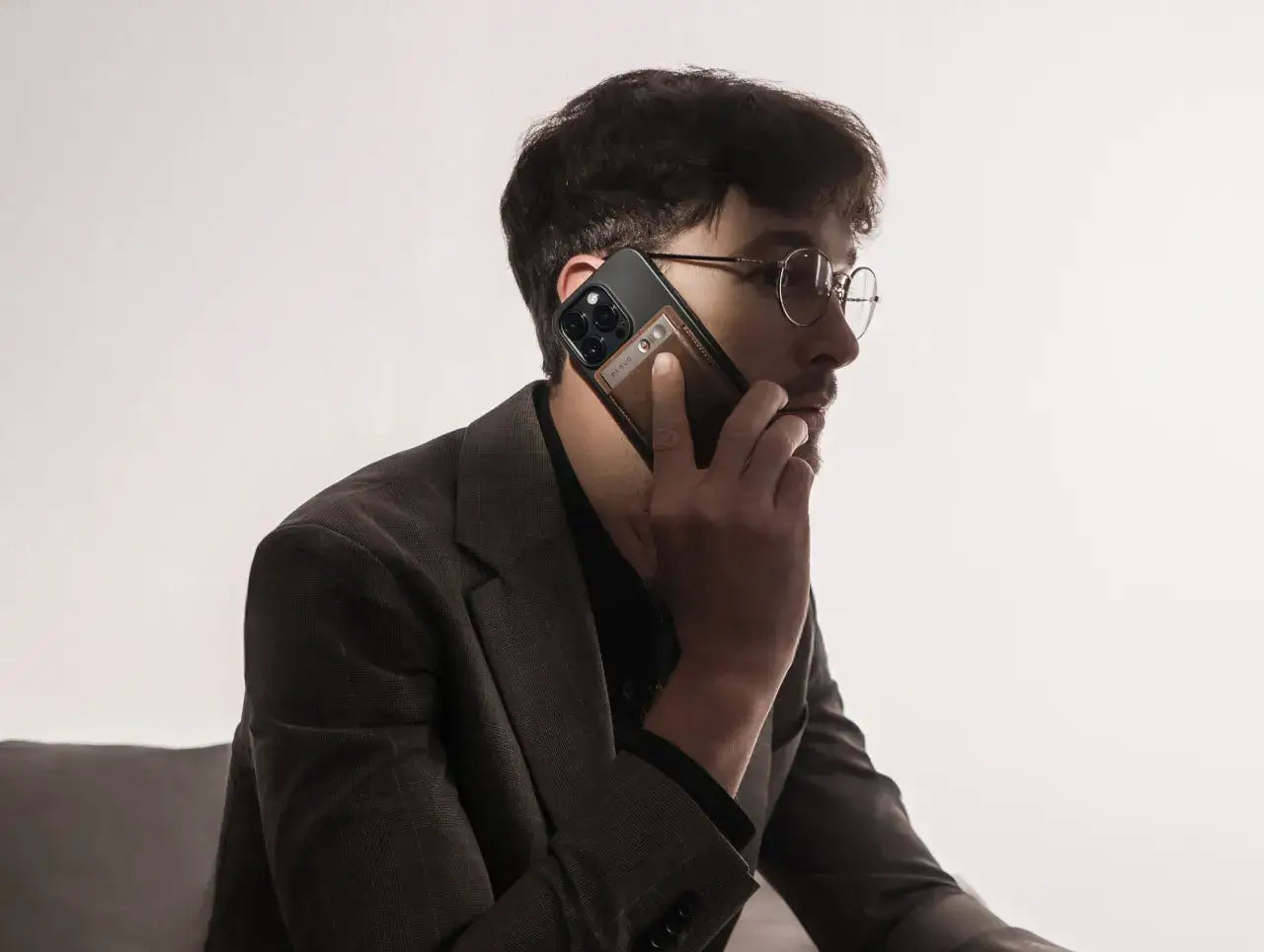 Man in a suit holding a smartphone with a silver Plaud Note in a brown protective case attached to the back for phone call recording.