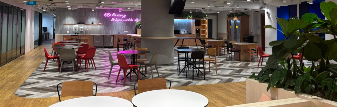 Modern office lounge with colorful chairs, herringbone floors, and a neon sign in a spacious break area.