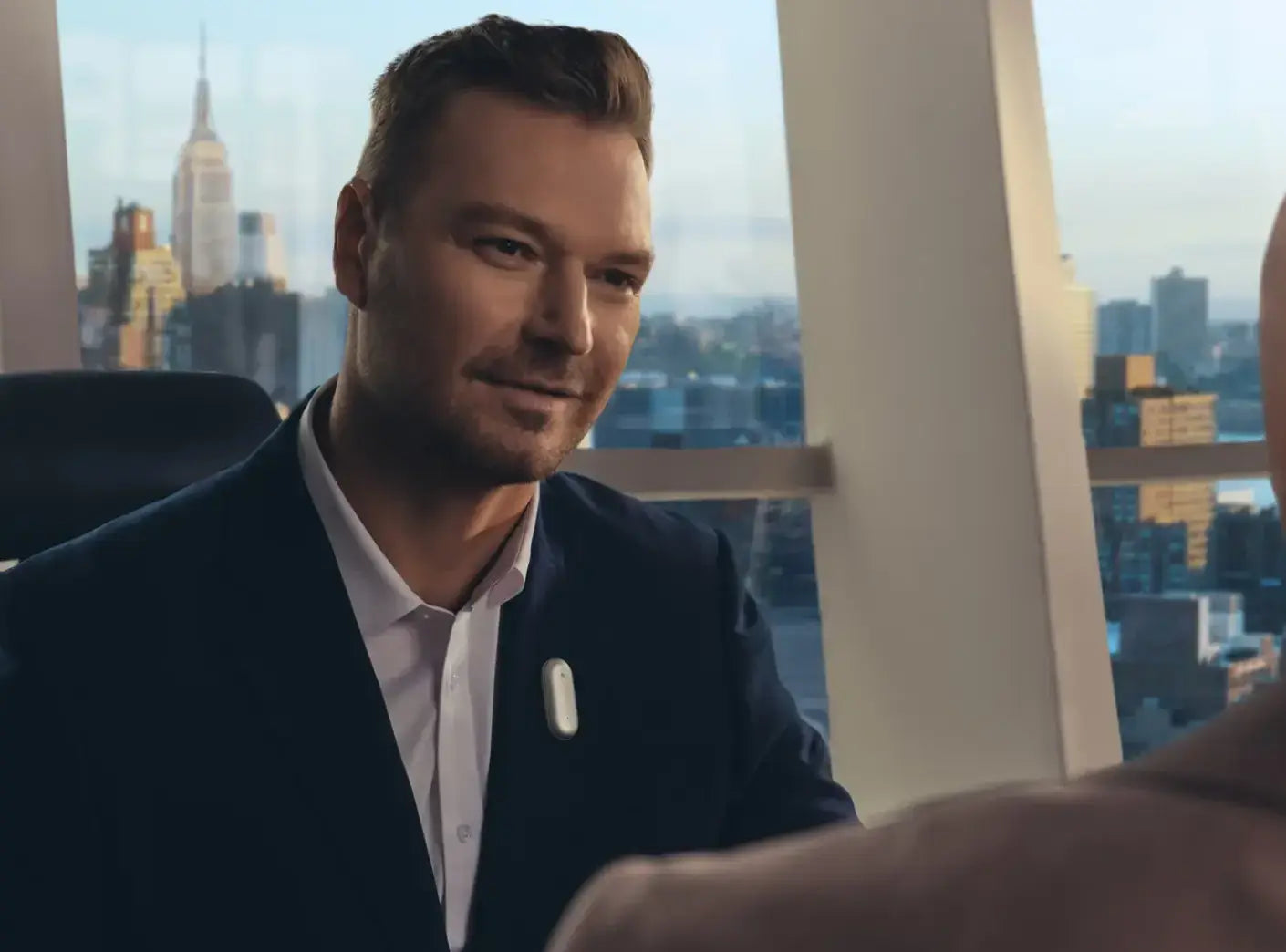 Man in a suit wearing a silver Plaud NotePin on his lapel during a meeting in a high-rise office.