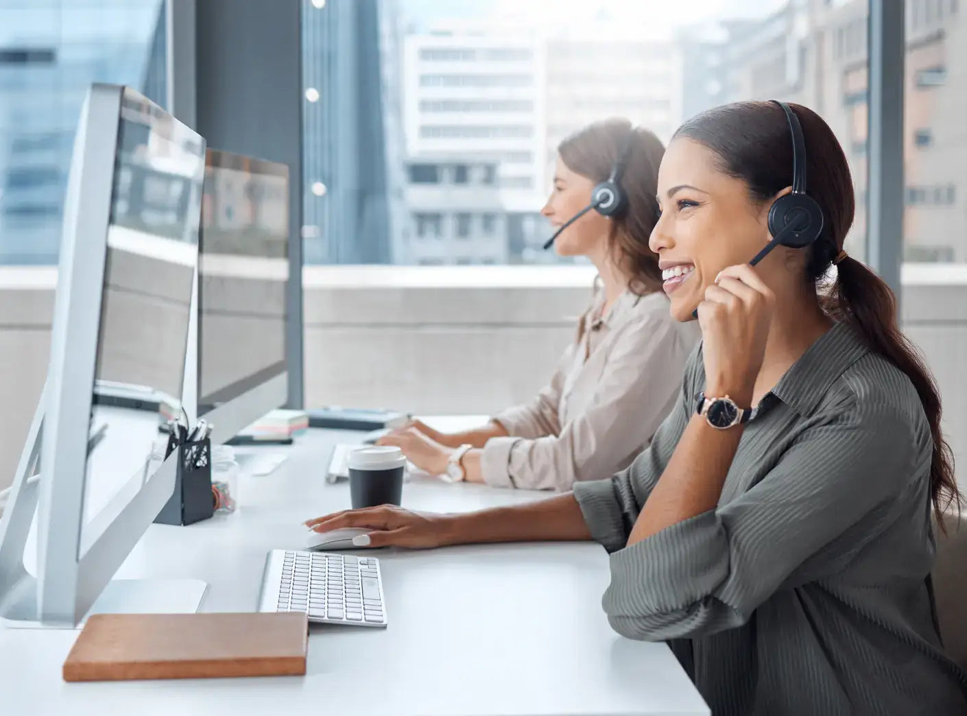 Two professional women wearing headsets work at desktop computers in a modern office setting.